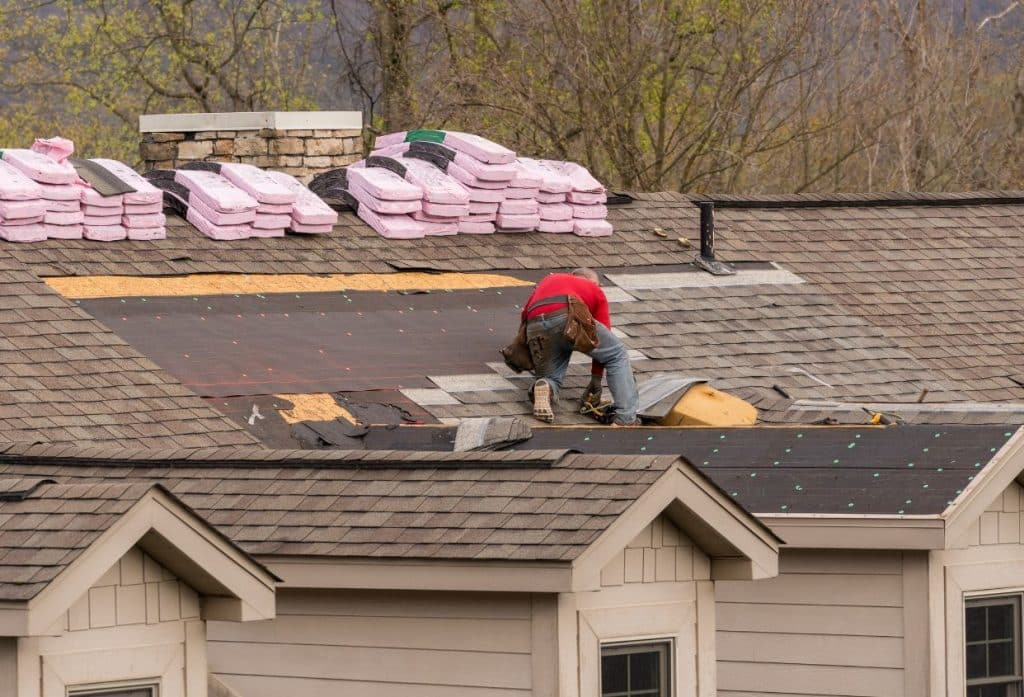 A roofer in a red shirt repairs a shingle roof, with exposed underlayment and stacks of pink roofing materials nearby.