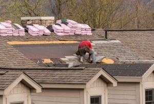 A roofer in a red shirt repairs a shingle roof, with exposed underlayment and stacks of pink roofing materials nearby.