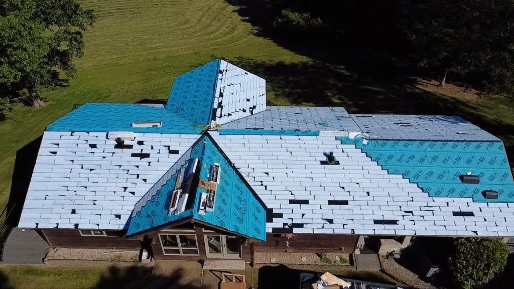 An aerial view shows a house undergoing roof replacement, with new shingles and blue protective underlayment covering sections of the roof.